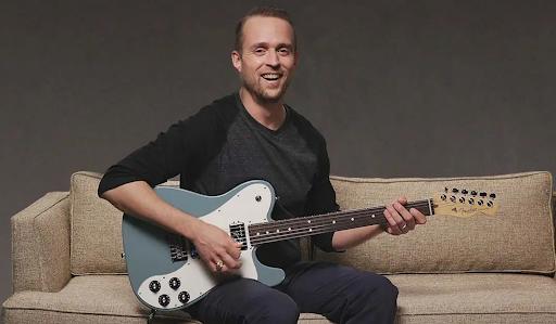 A smiling guitar player seated on a couch with a blue electric guitar