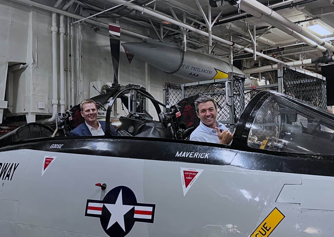 Tom Cronkright, left, and Tyler Adams, right, smiling in an aircraft cockpit labeled Goose and Maverick.