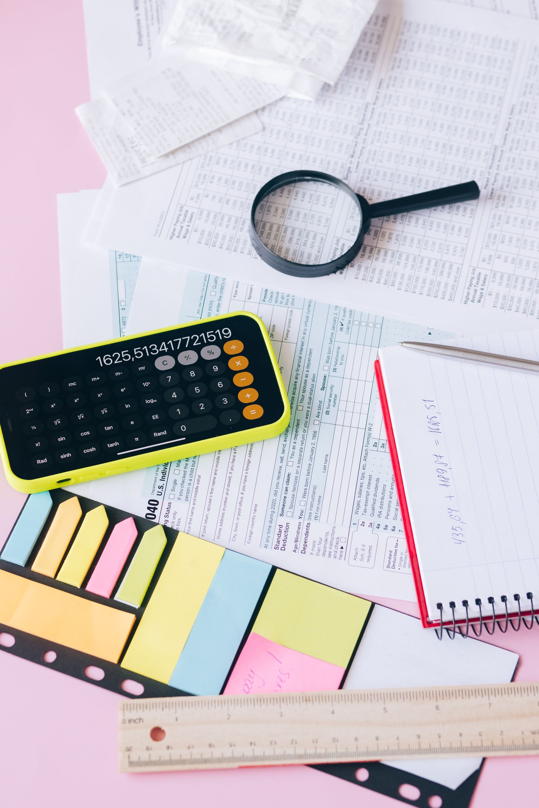 A calculator and magnifying glass beside paperwork on a pink desk.