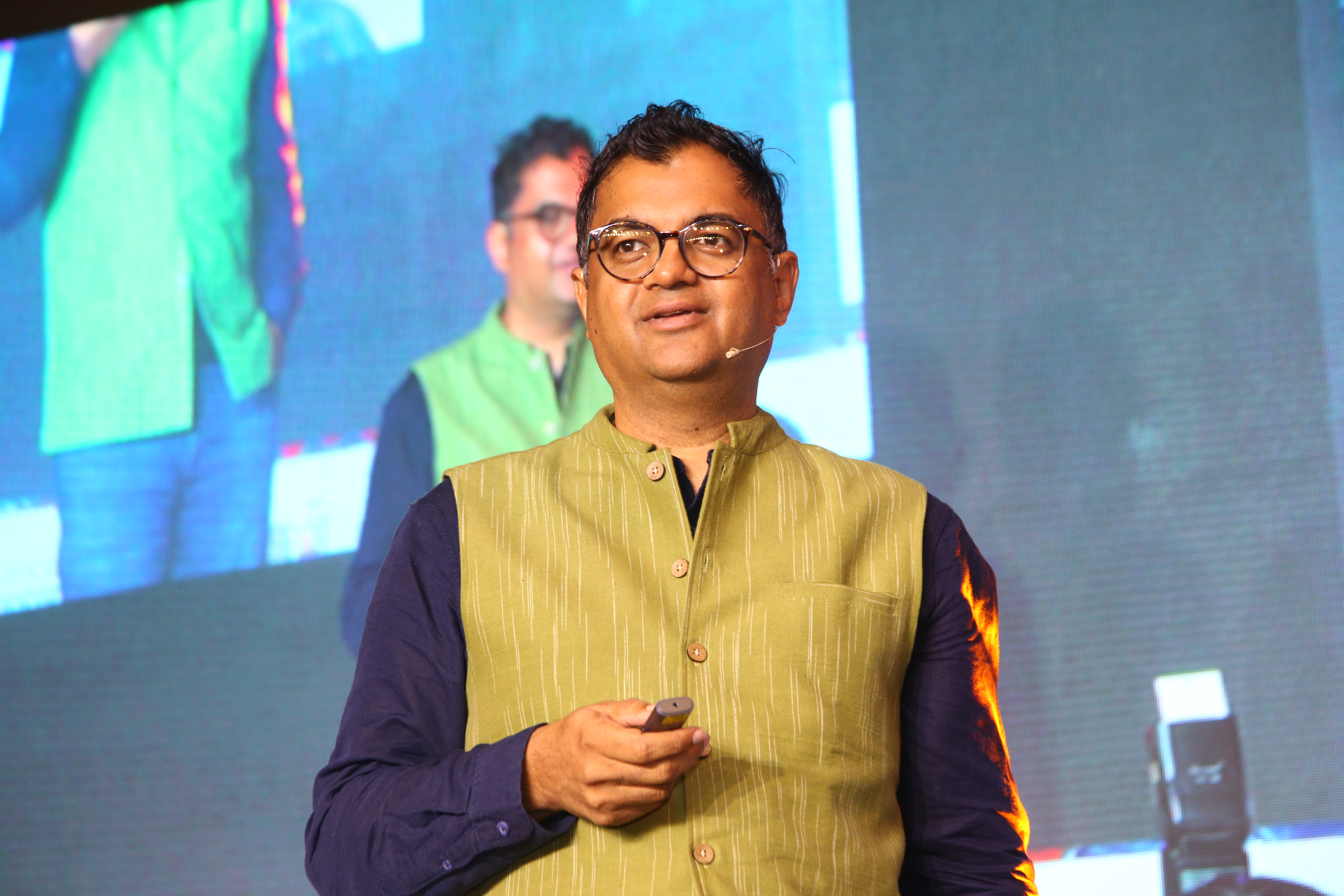 Umakant Soni on stage wearing glasses, a yellow-green waistcoat and a headset microphone