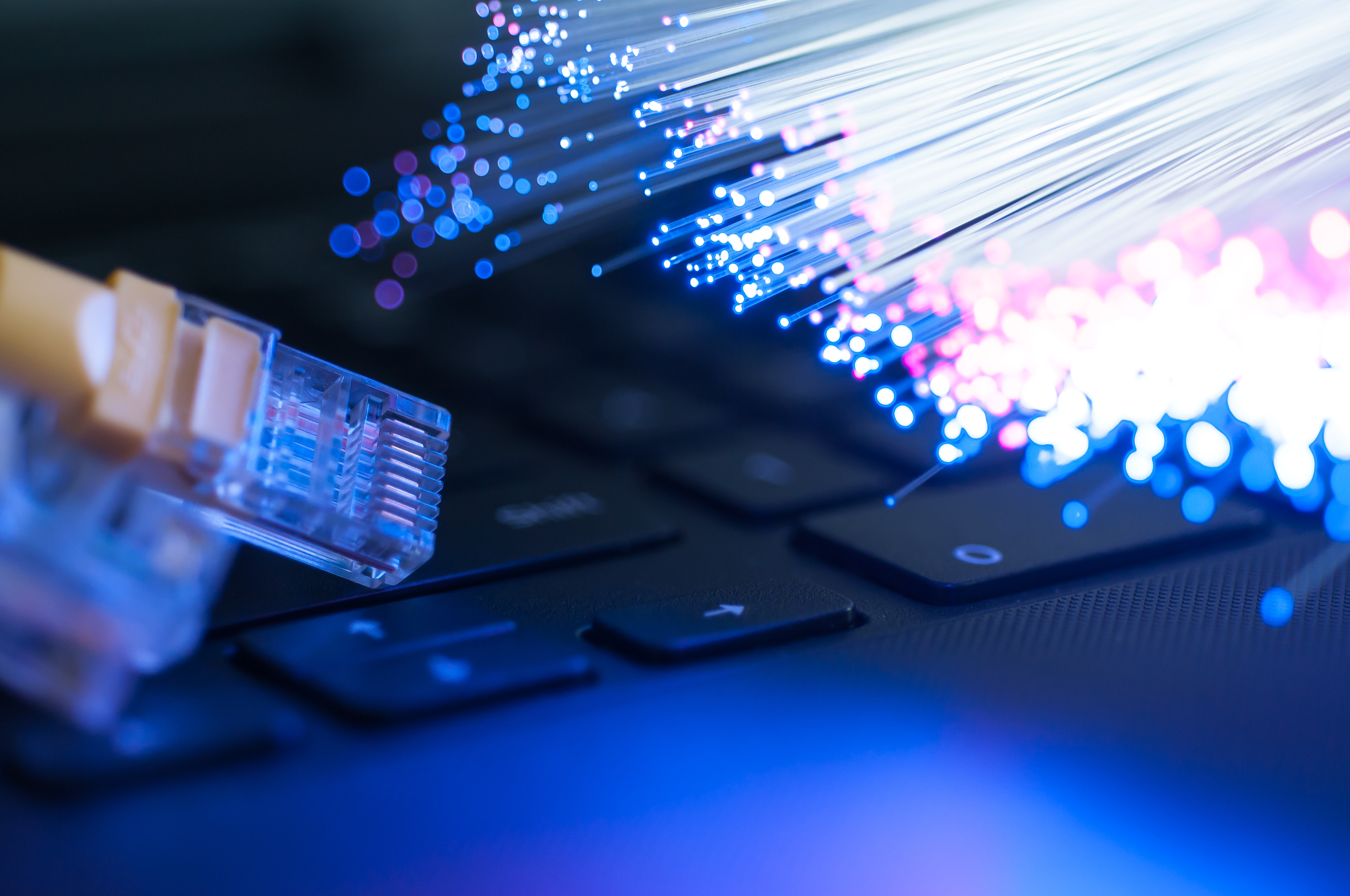 Close-up illustrative photograph of a yellow Ethernet cable beside illuminated optical fibres and a laptop keyboard