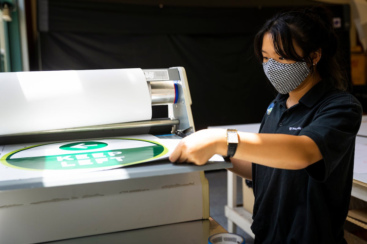A masked University Graphic Systems student laminating a green Keep Left campus safety sign