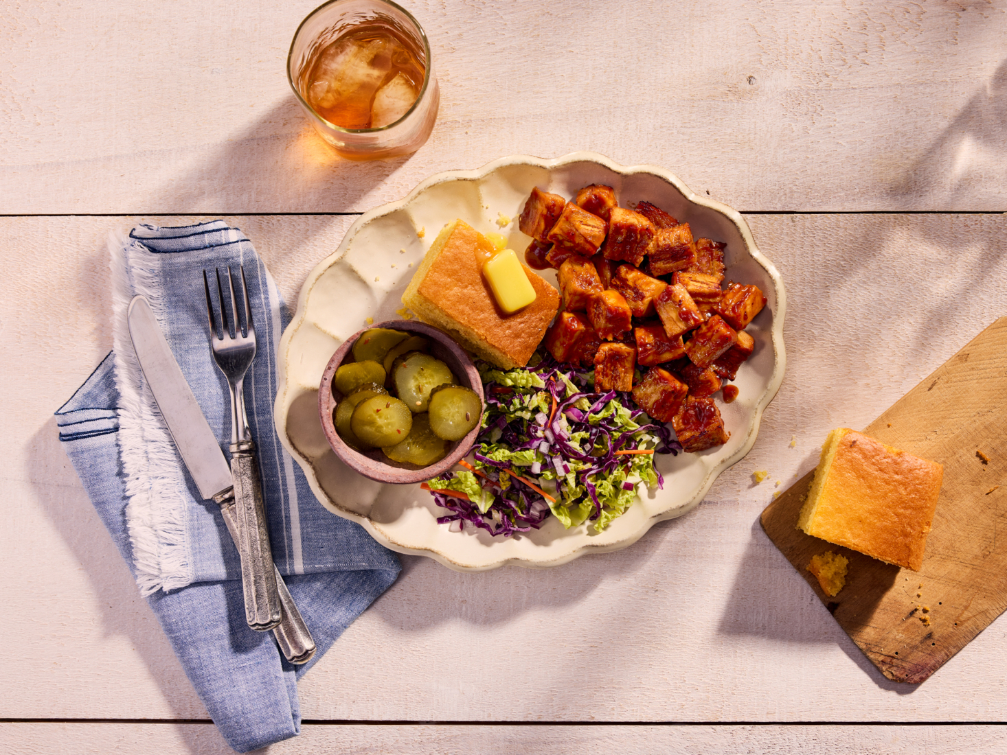 A plated barbecue cultivated chicken dish with cornbread, slaw and pickles on a rustic table