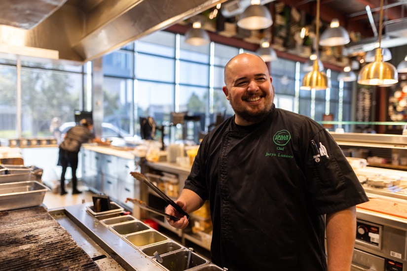 An Urban Plates chef stands with tongs at a kitchen counter