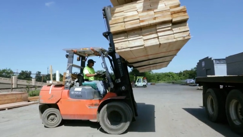 A forklift lifts a stack of lumber beside a flatbed truck at a US LBM operation