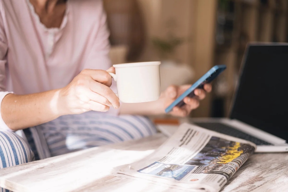 A reader holds a coffee and smartphone beside an open newspaper and laptop