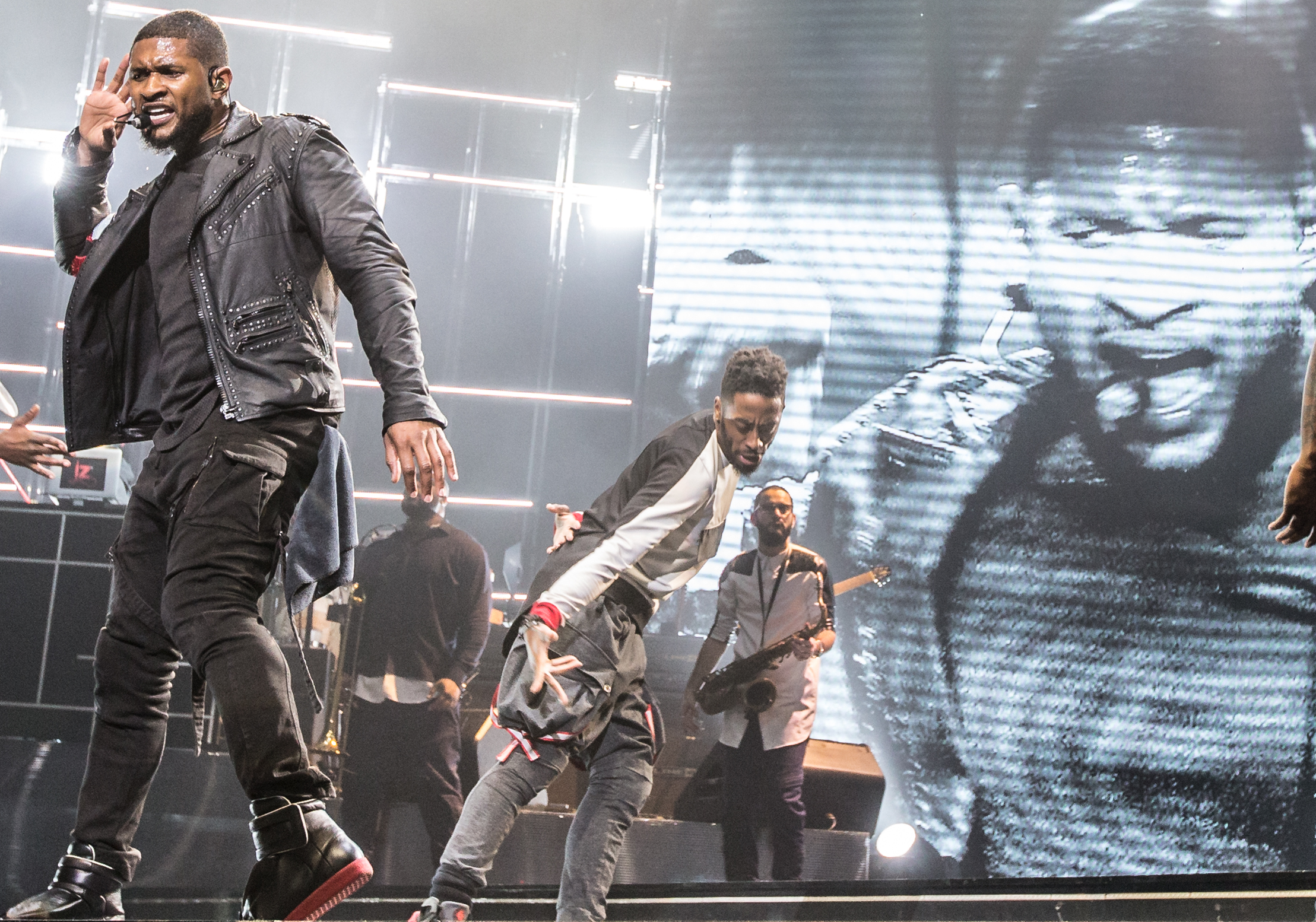 Usher performs on an arena stage in dark clothing and sunglasses, one hand raised toward the audience
