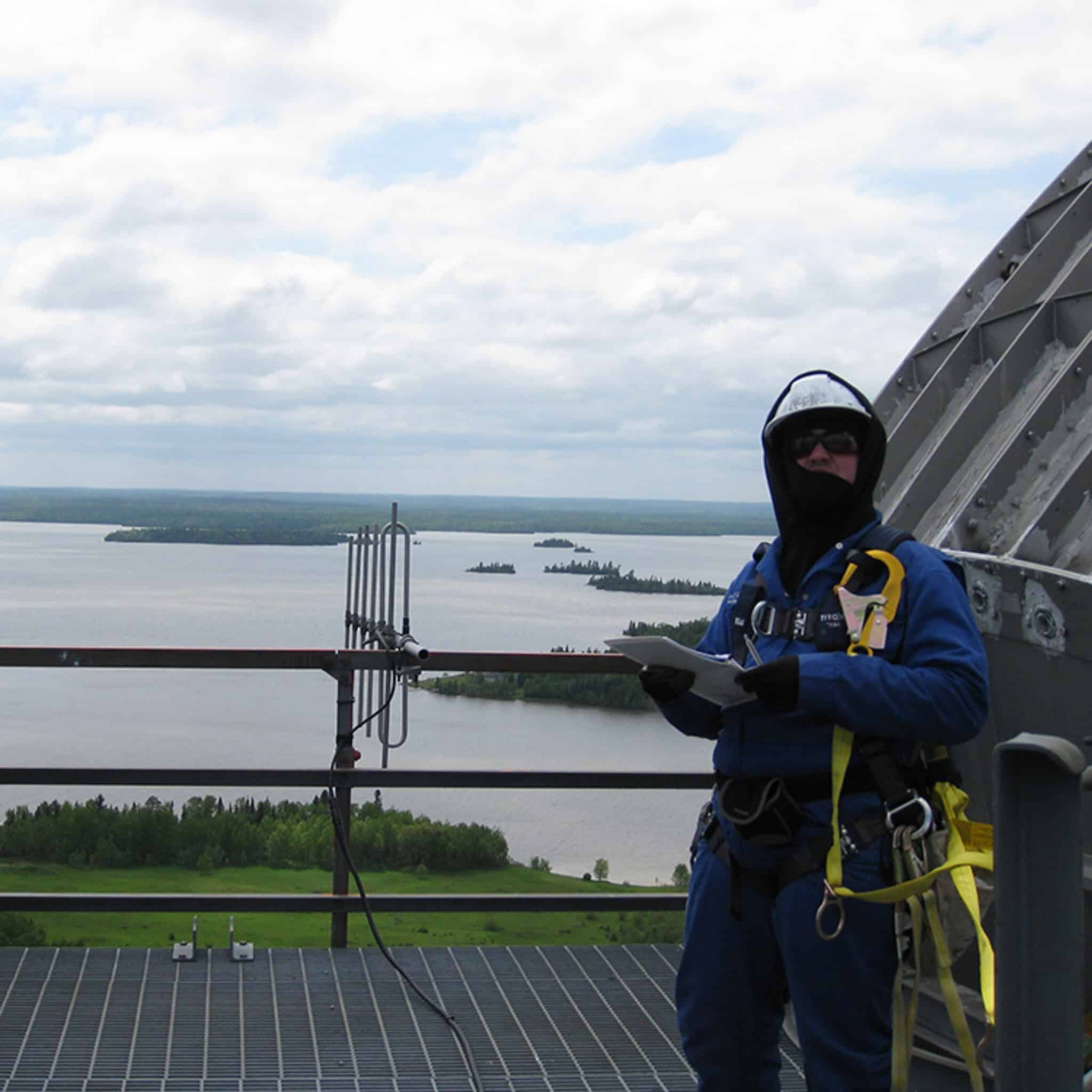 A harnessed tower inspector holding a clipboard high above a lake