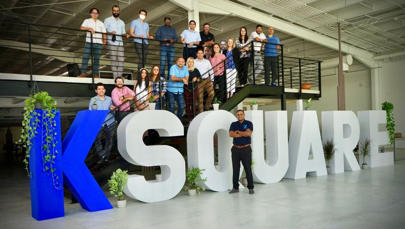 Varma Buddharaju stands beside Ksquare lettering with colleagues gathered on the staircase behind him