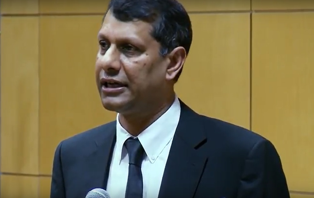 Venkat Srinivasan holding a microphone while speaking, wearing a dark suit and tie