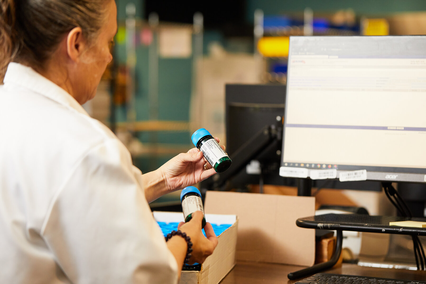 A Vetsource pharmacy staff member checks medication bottles beside a computer before packing