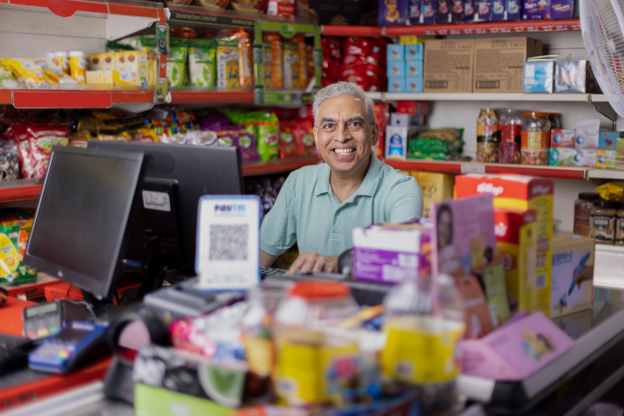 A smiling man behind a shop counter surrounded by groceries and a computer