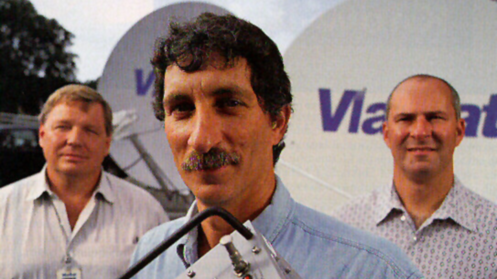 Archival Viasat team photograph with Mark Dankberg in the foreground and satellite dishes behind the group