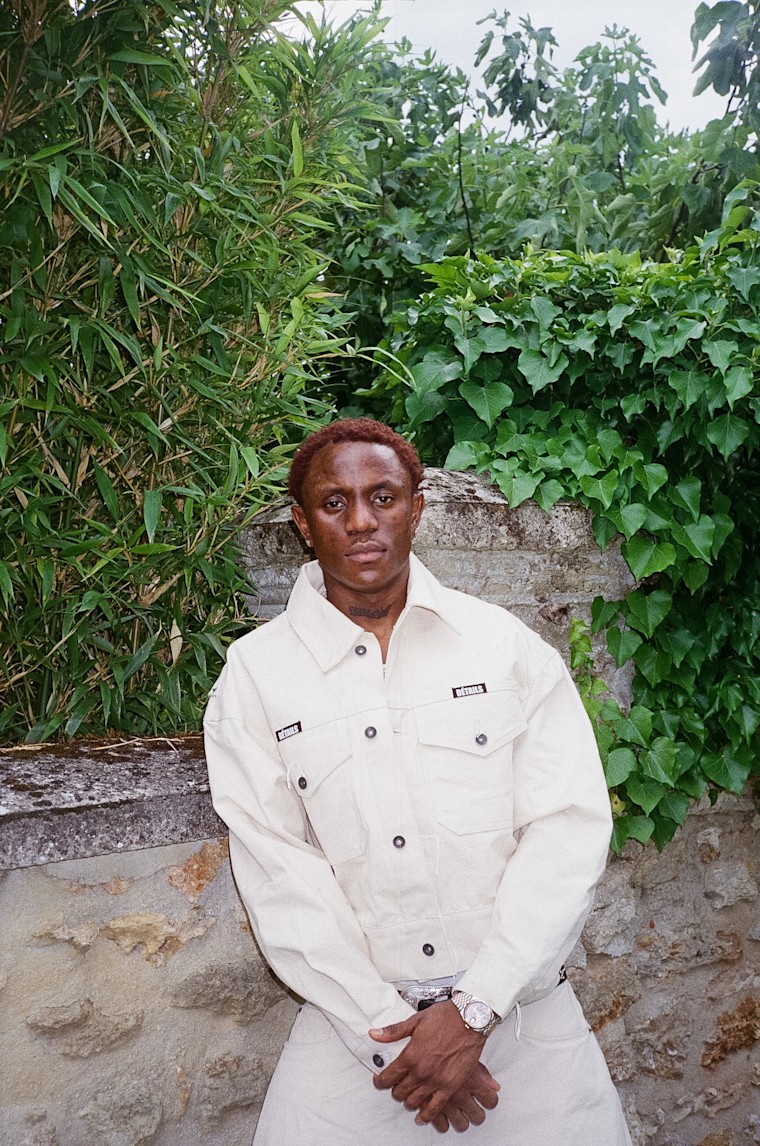 Victony in a cream jacket standing against an ivy-covered stone wall