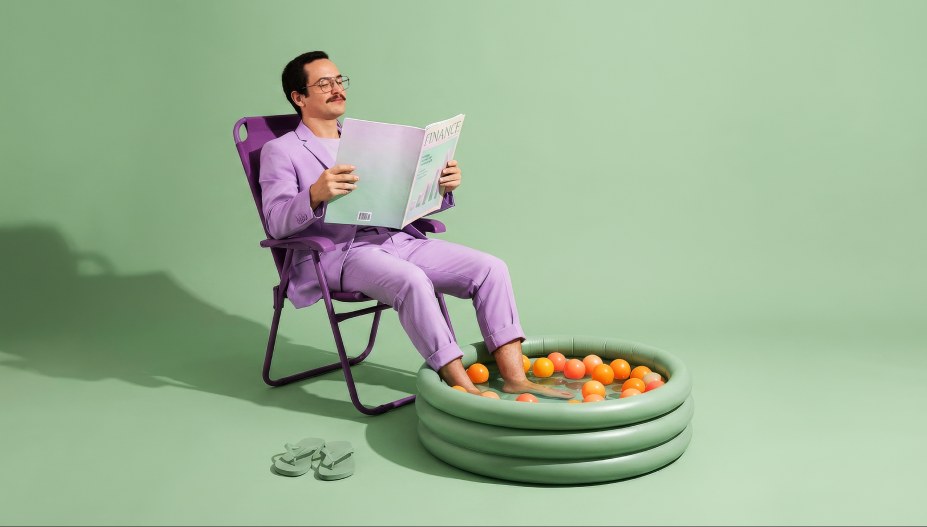 A man in a lilac suit relaxes in a folding chair reading a magazine titled Finance, feet in a small inflatable pool