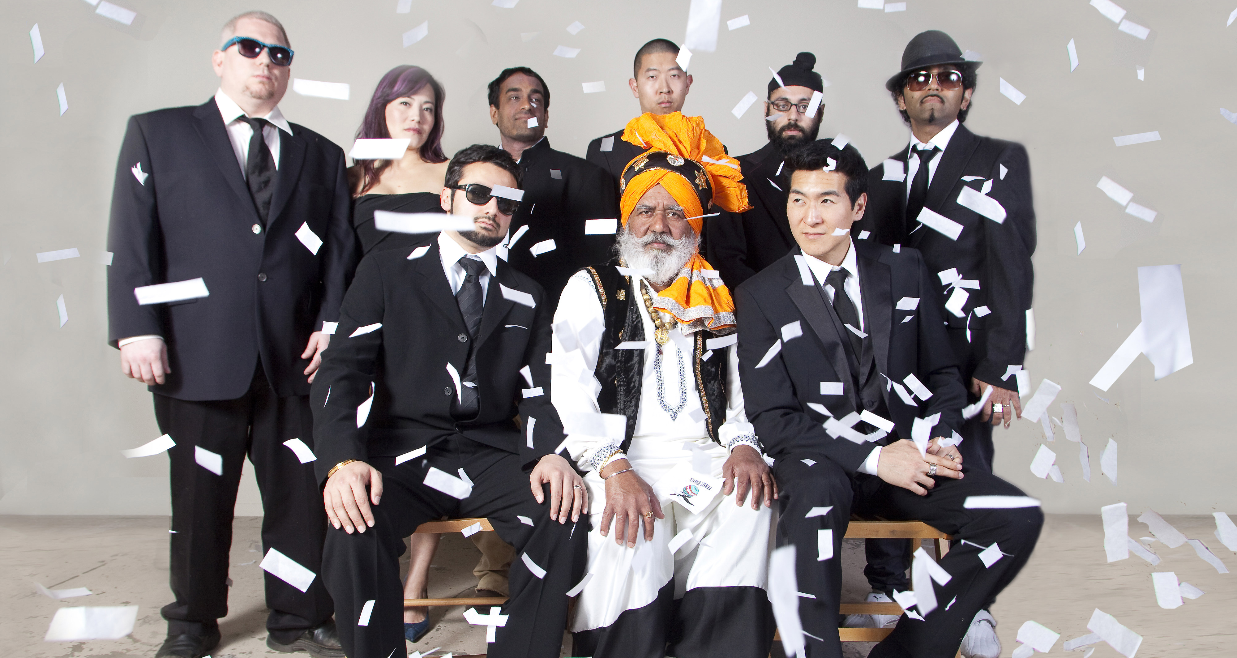 BlackMahal band members in dark suits, with Ustad Lal Singh Bhatti seated at the center in an orange turban, surrounded by falling confetti.