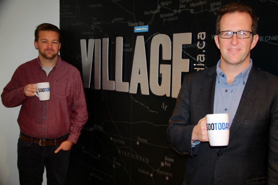 Village Media executives Jeff Elgie and Jake Cormier holding SooToday mugs in front of a Village Media wall map