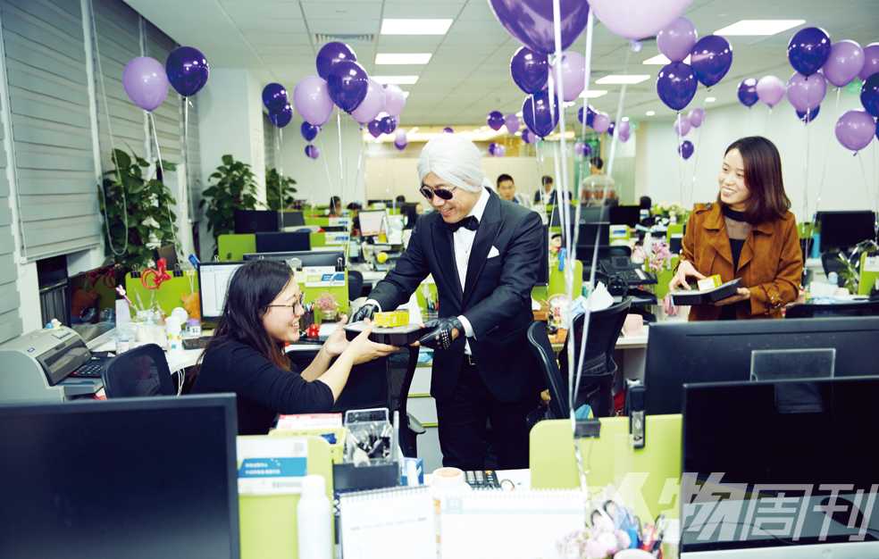 A suited figure wearing a white wig and sunglasses serves cake to a colleague in a balloon-filled Baozun office
