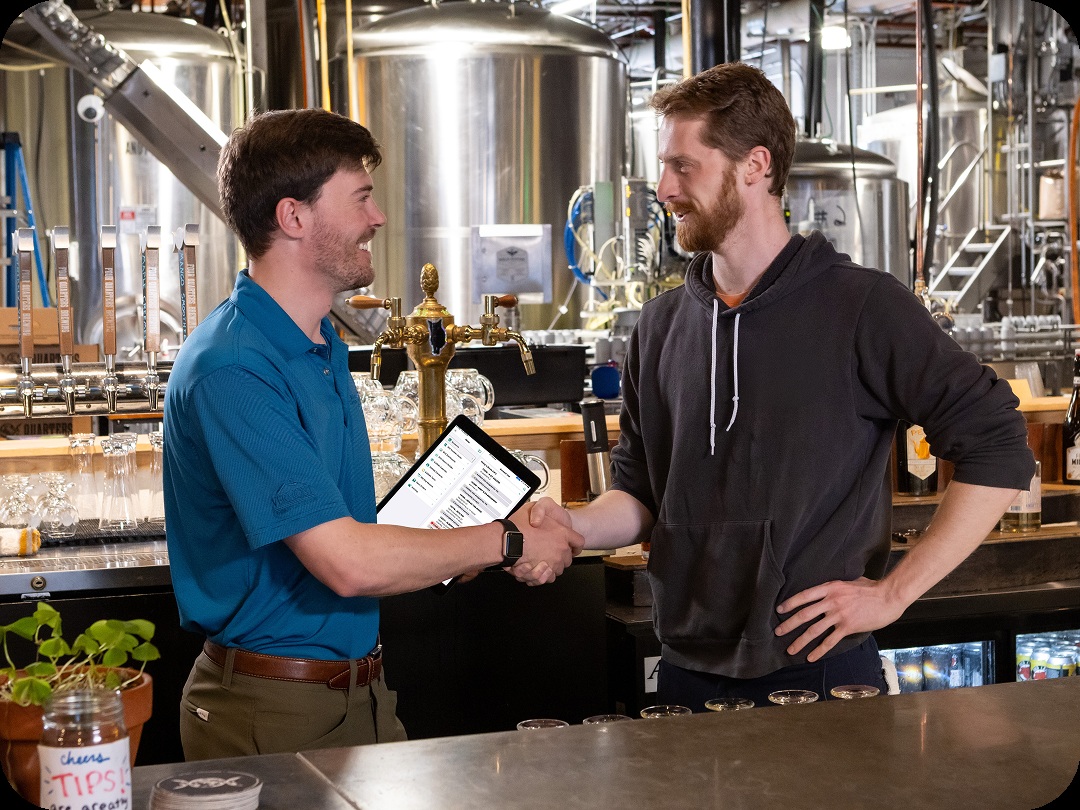 Two people shake hands across a brewery bar, with a tablet between them