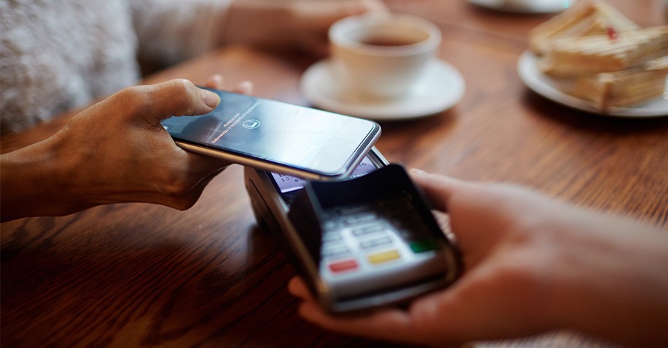 A customer holds a smartphone over a payment terminal at a cafe table.