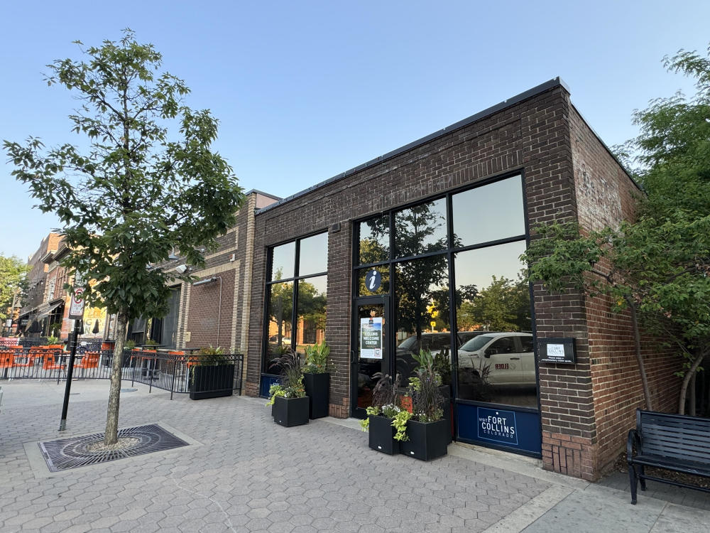 The brick Visit Fort Collins Welcome Center in Old Town Square, with large windows and planters by the entrance