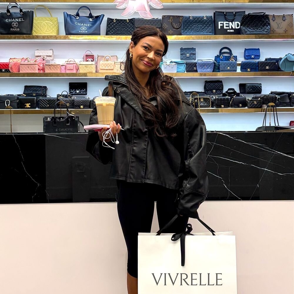 A visitor holding a shopping bag and iced drink in front of Vivrelle’s designer handbag shelves