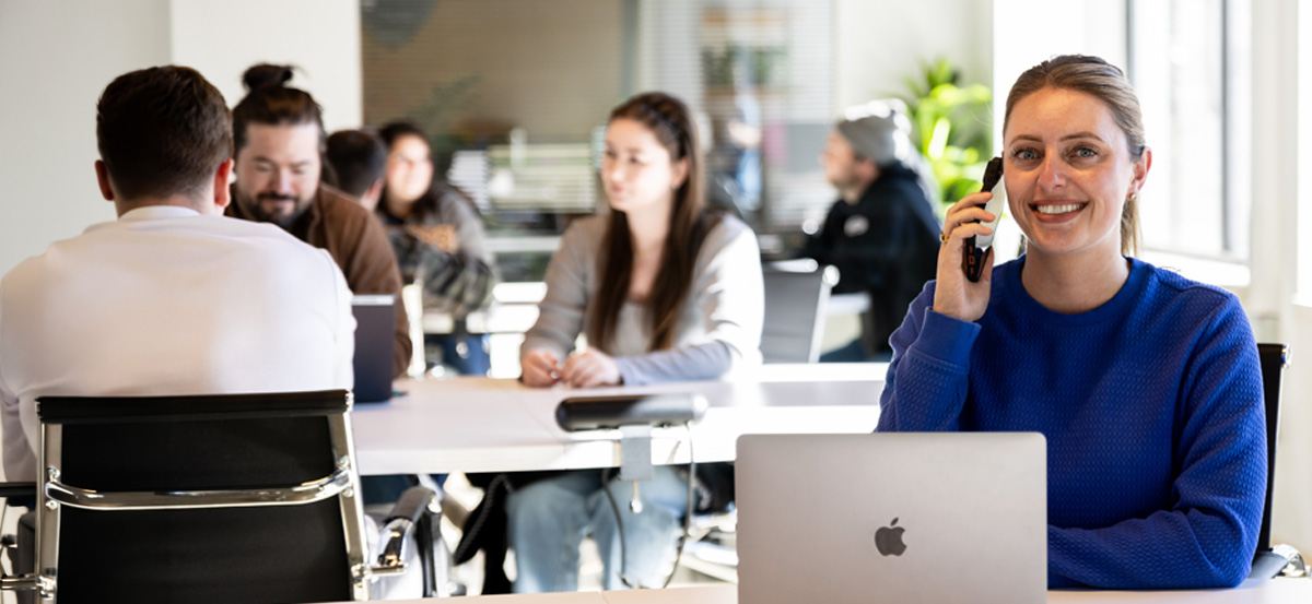 VOIP.COM promotional photograph showing a woman on a phone beside a laptop in a shared workspace
