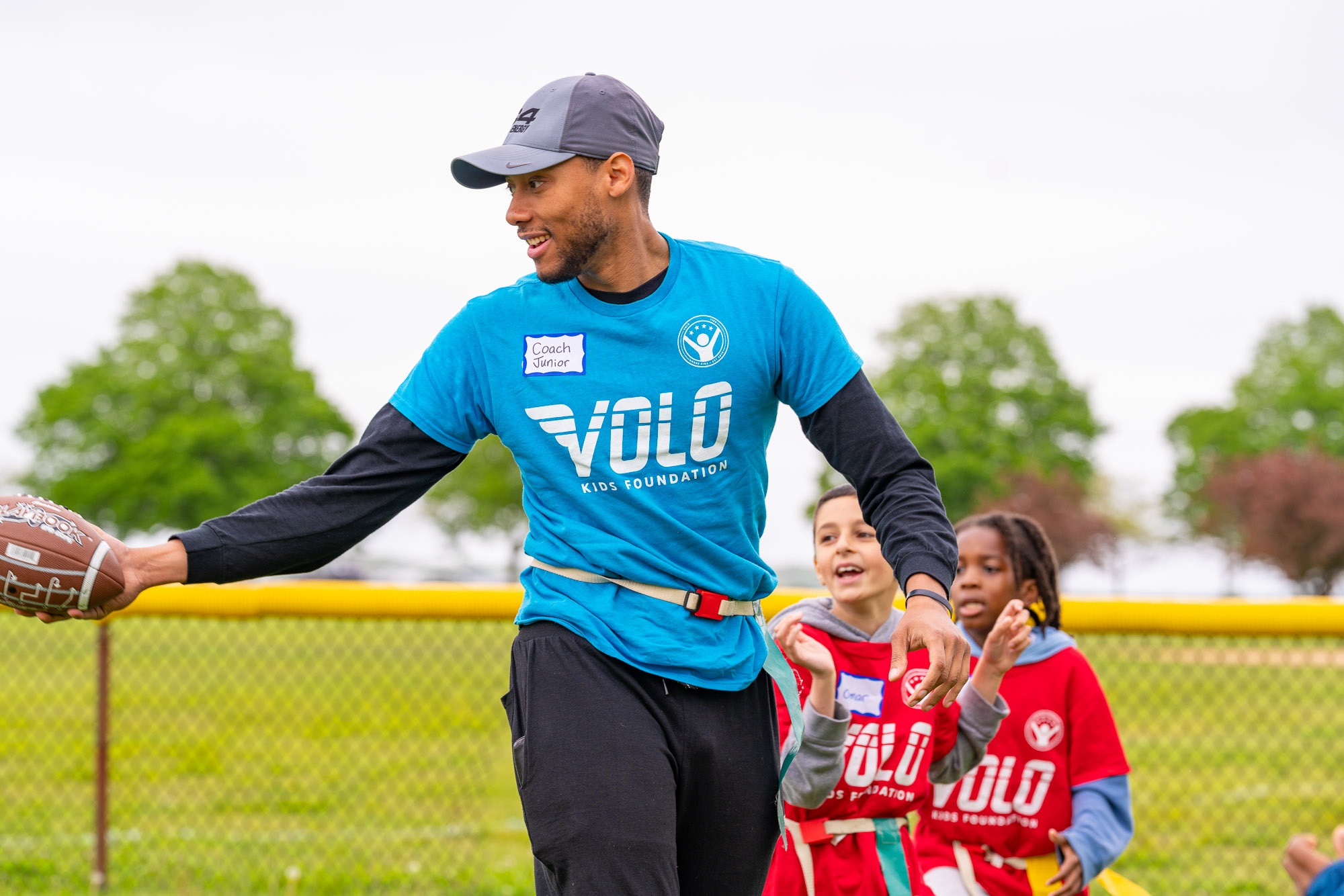 A Volo Kids coach holding a football as children play behind him