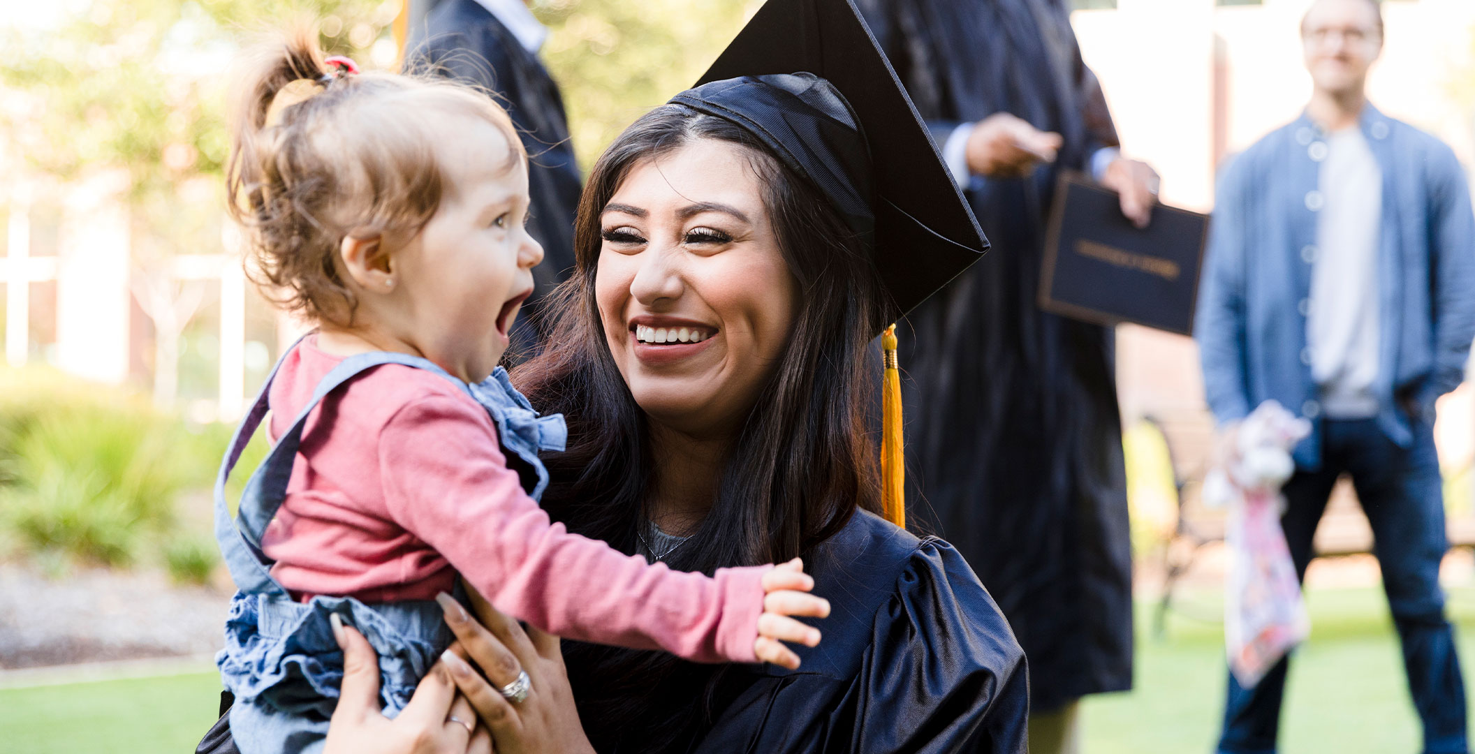 An adult college graduate holding a young child at commencement