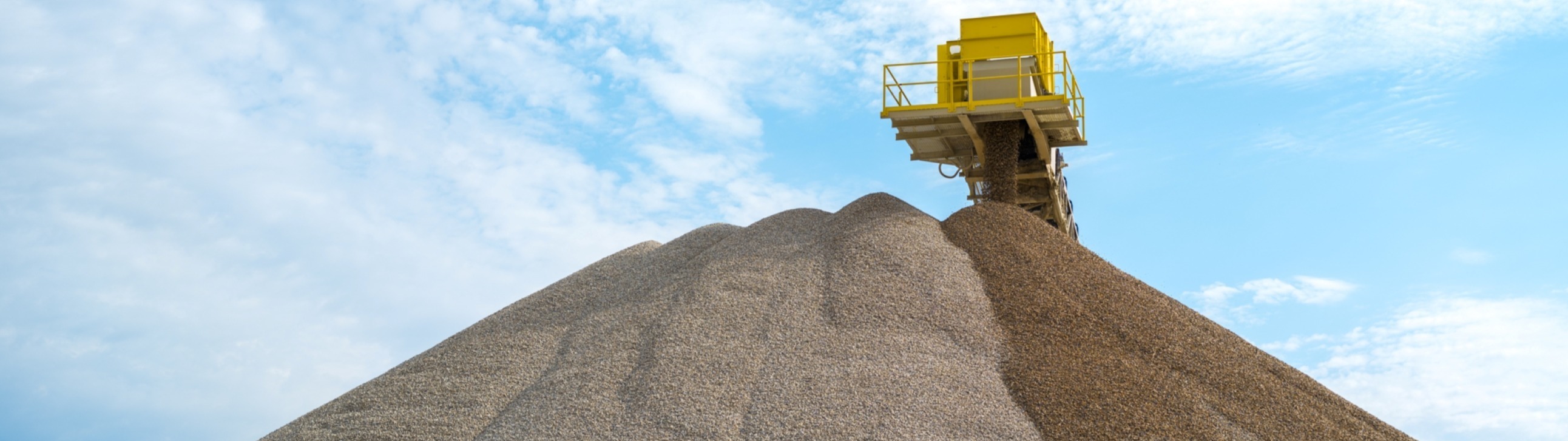 An elevated conveyor builds aggregate stockpiles beneath a blue sky