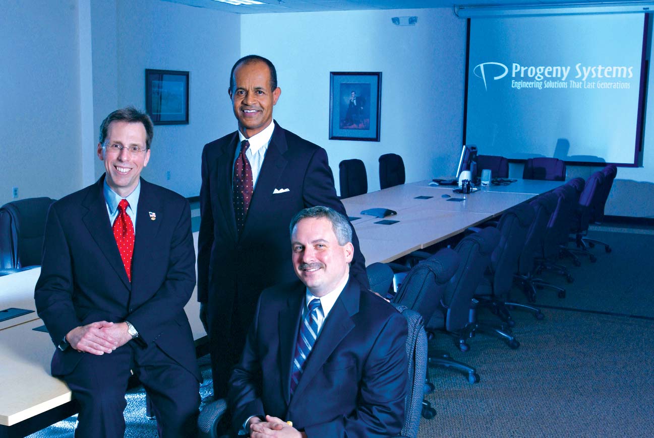 Chris Bergstrom and Seth Carter standing beside Walter P. Kitonis III, seated in a Progeny Systems conference room
