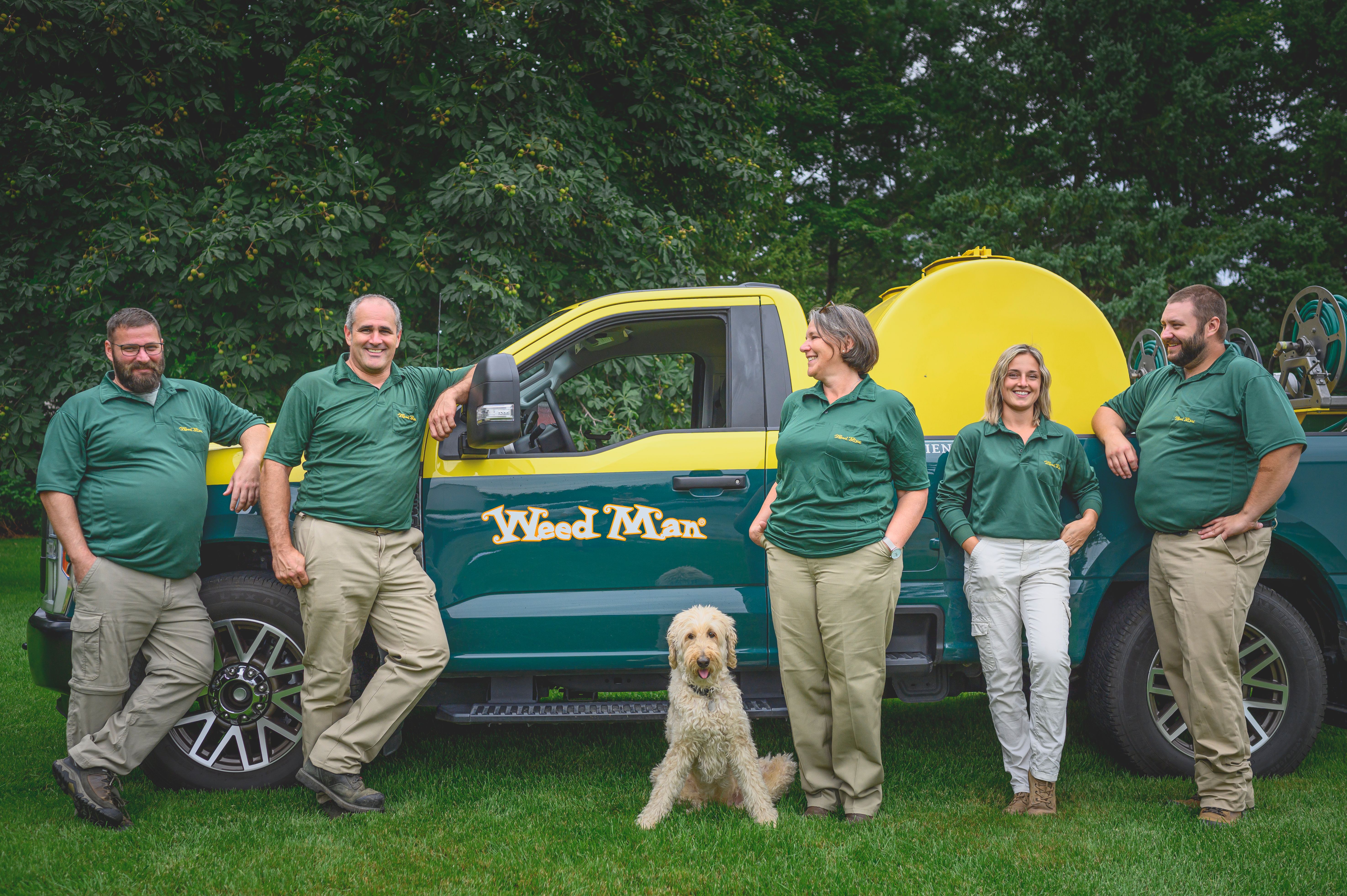 Five Weed Man lawn-care professionals beside a green and yellow service truck with a dog seated on the lawn