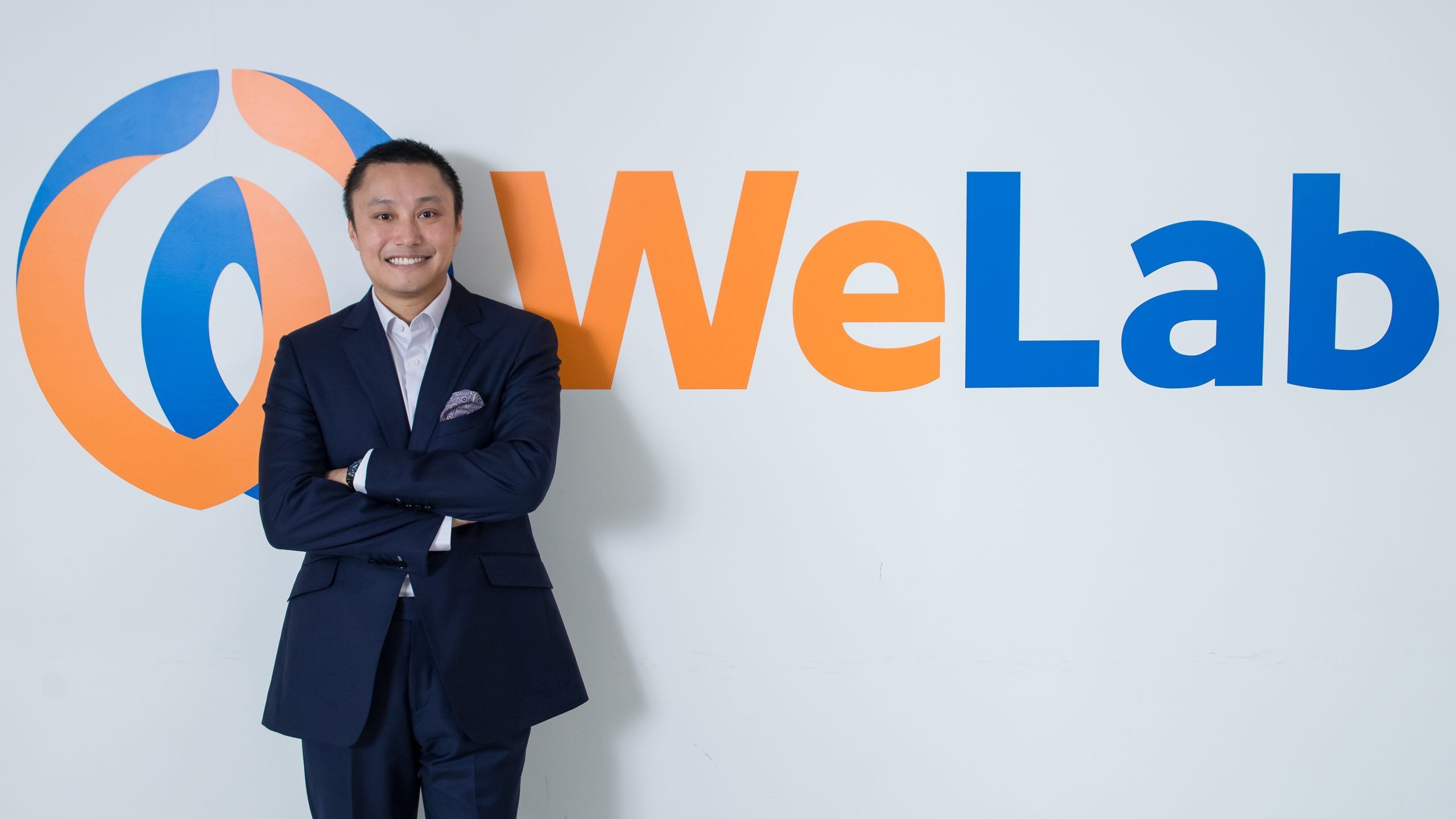 WeLab co-founder Simon Loong standing with arms folded beside the company’s orange and blue wall sign