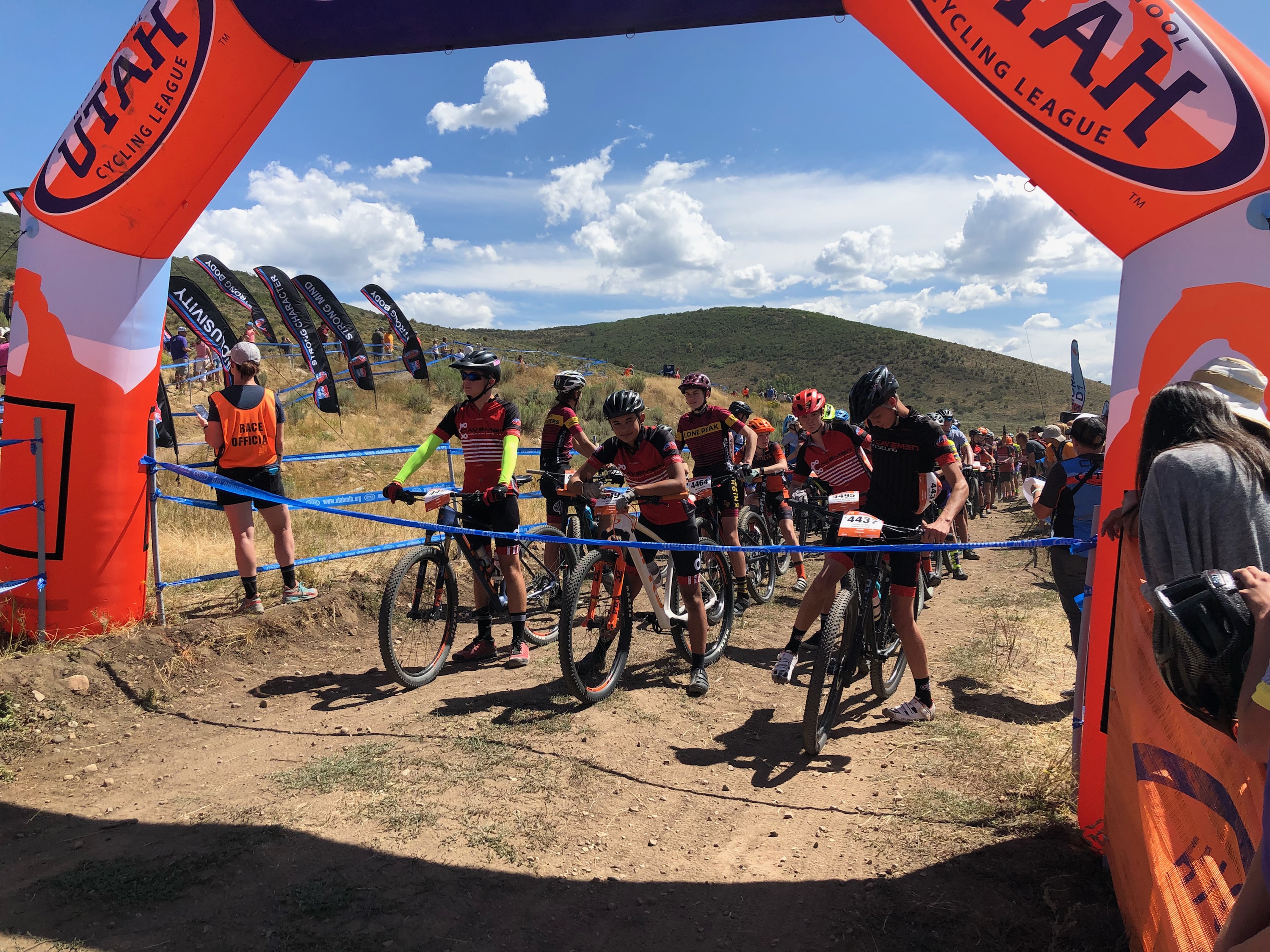 Young cyclists waiting beneath an orange Utah cycling league arch at an outdoor event