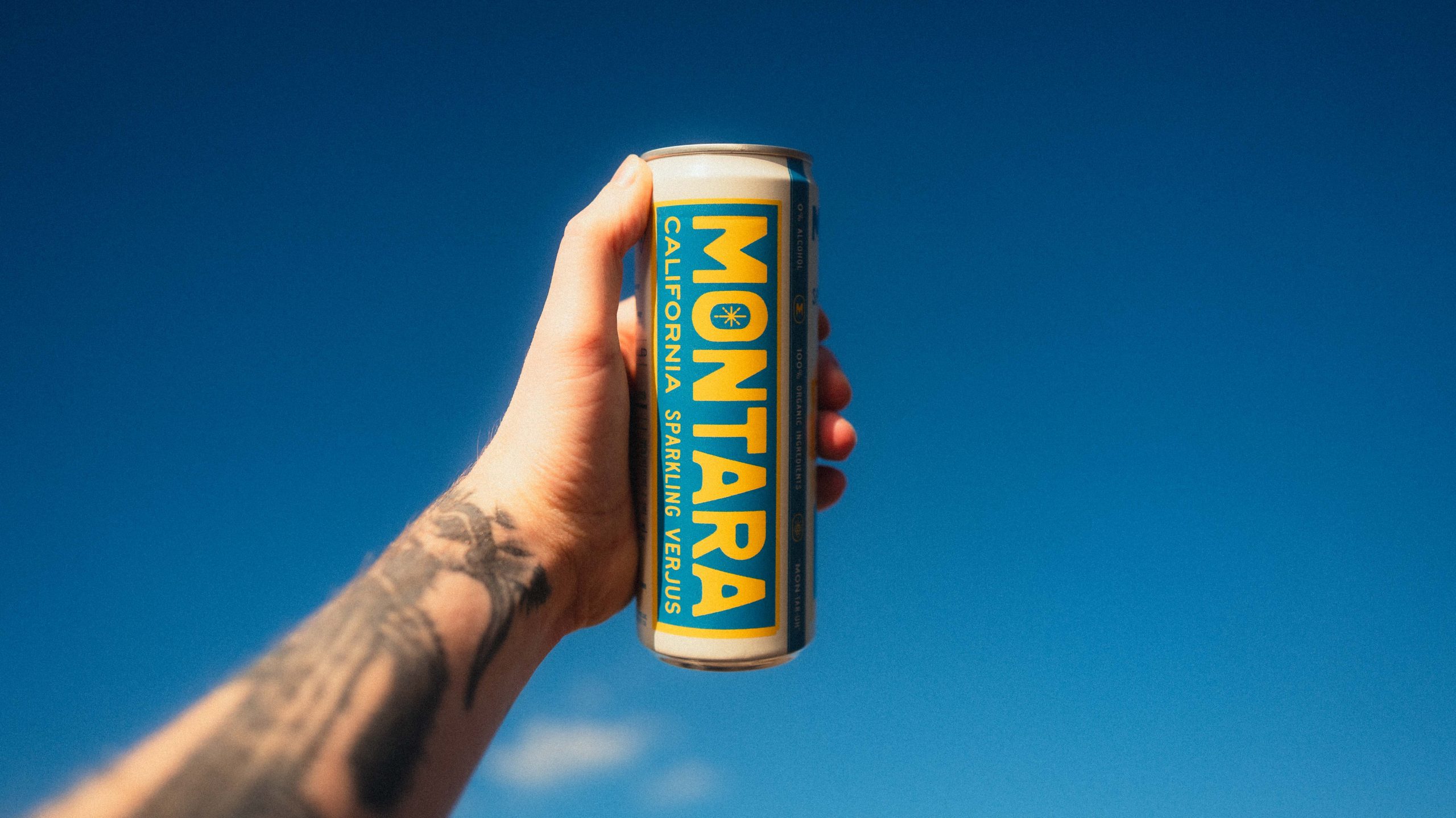 A hand holds a yellow-and-blue Montara sparkling verjus can against a blue sky