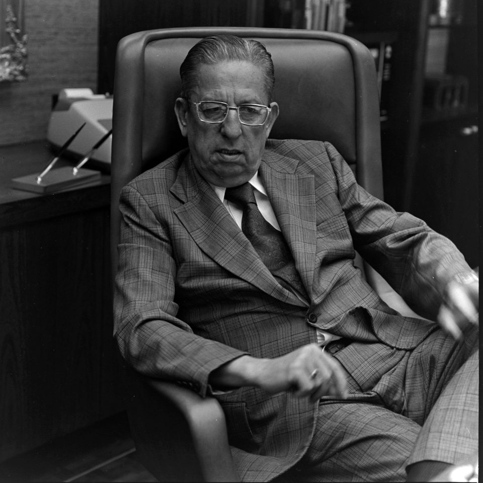 William T. Dillard seated in a leather office chair at his desk, wearing a checked suit and glasses.