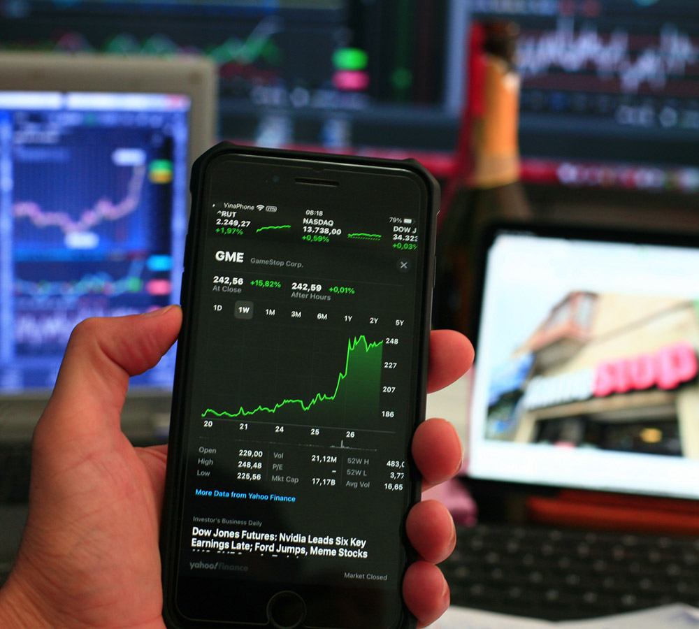 A hand holds a phone showing GameStop market data in front of several trading screens