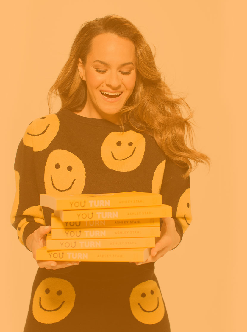 Ashley Stahl smiling while holding a stack of her You Turn books, in a yellow-toned promotional portrait