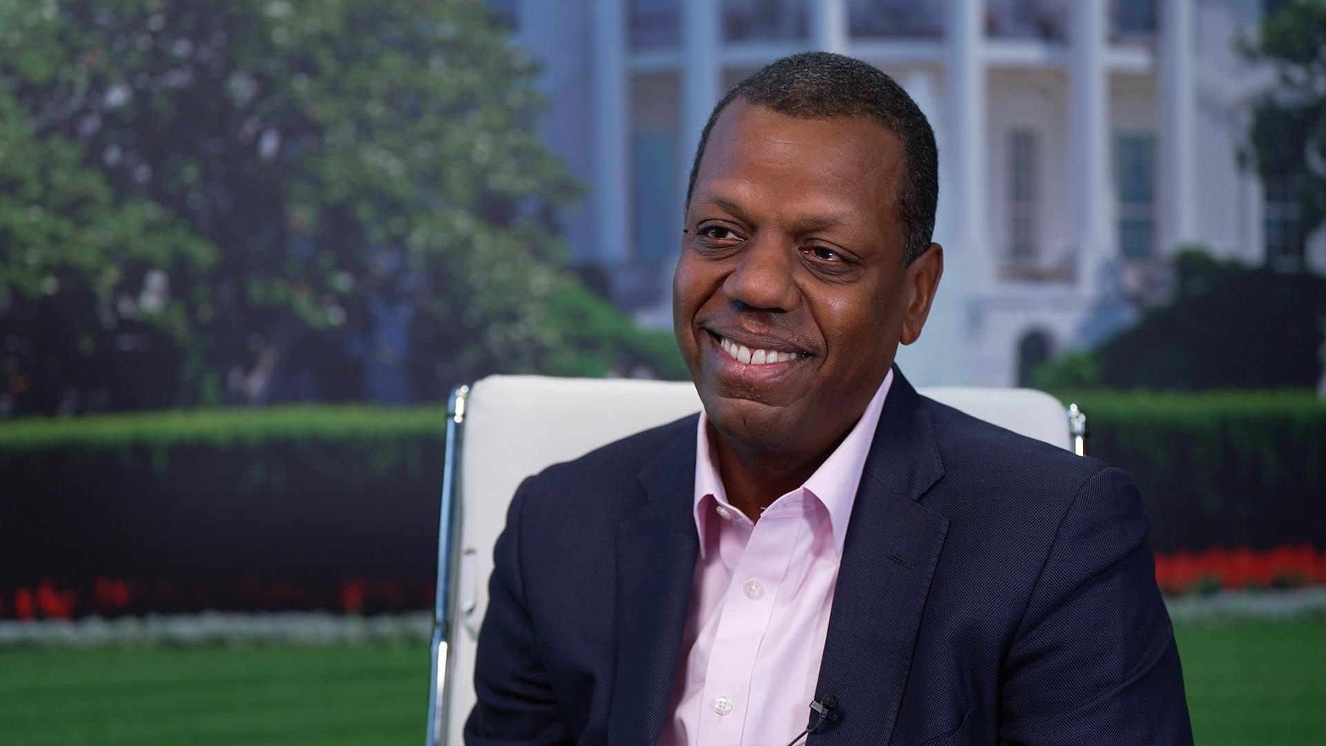 Wole Coaxum speaking on The Chavis Chronicles in a navy jacket and pink shirt