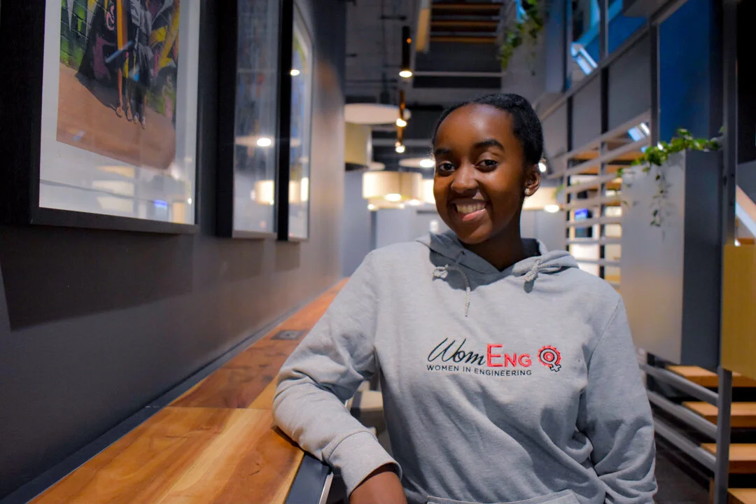 A smiling woman wearing a WomEng sweatshirt, seated at a table
