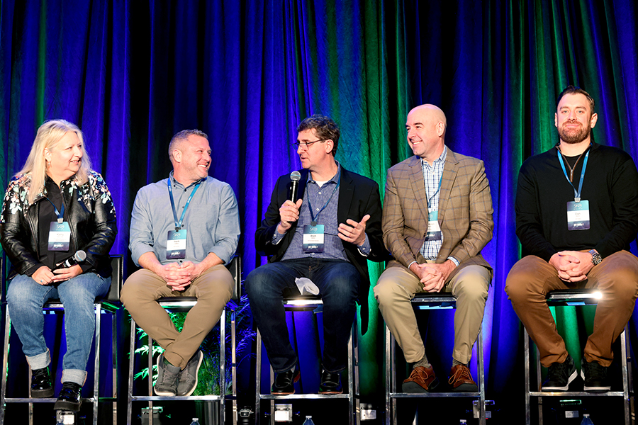 Five conference panelists seated onstage, with one speaking into a microphone