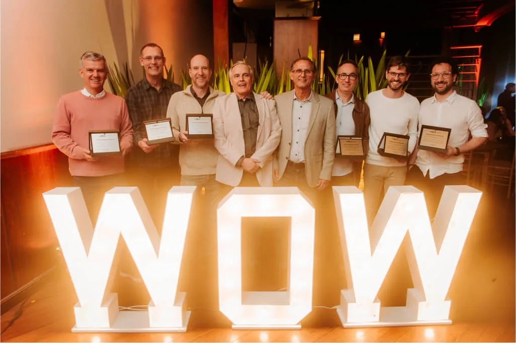 Eight people holding commemorative plaques behind illuminated WOW letters at a company event