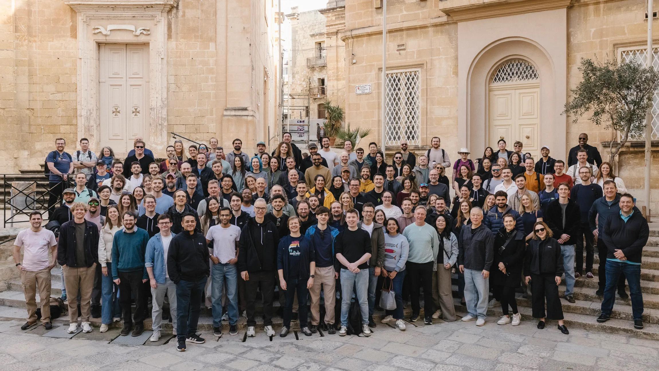 A large group of XBOW employees gathered outdoors in front of stone buildings