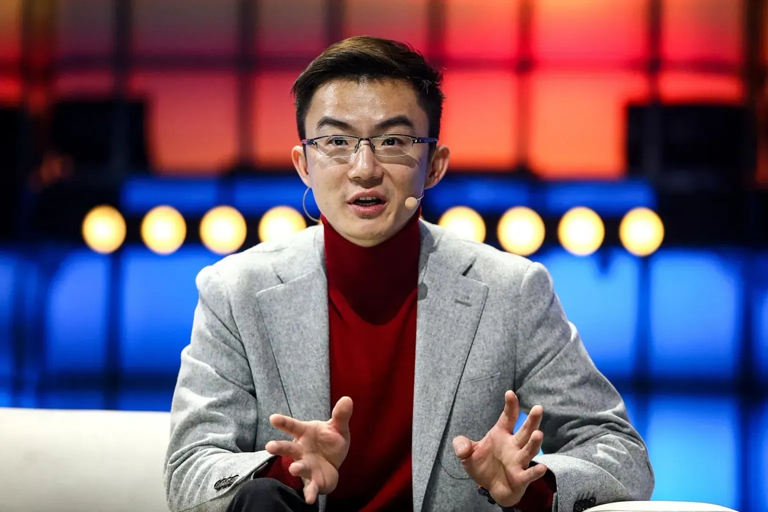 Xiaodi Hou speaking onstage in a gray jacket and red turtleneck at Web Summit in 2019