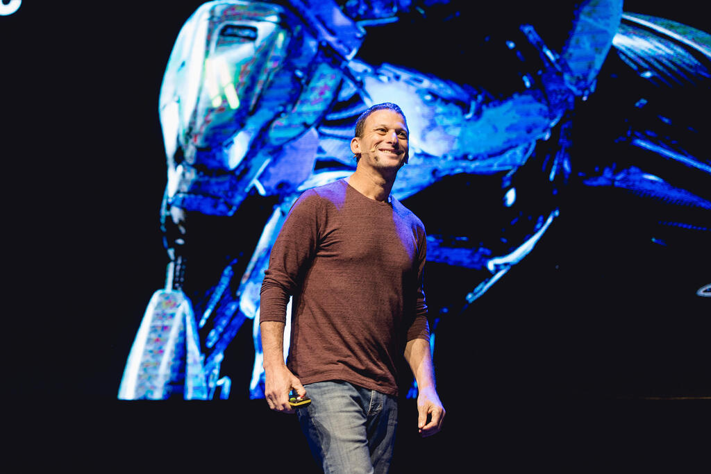 Yaki Faitelson onstage, holding a presentation remote, with a blue robot graphic behind him