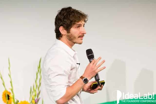 Yali Saar speaking onstage at an IdeaLab event, holding a microphone and presentation remote