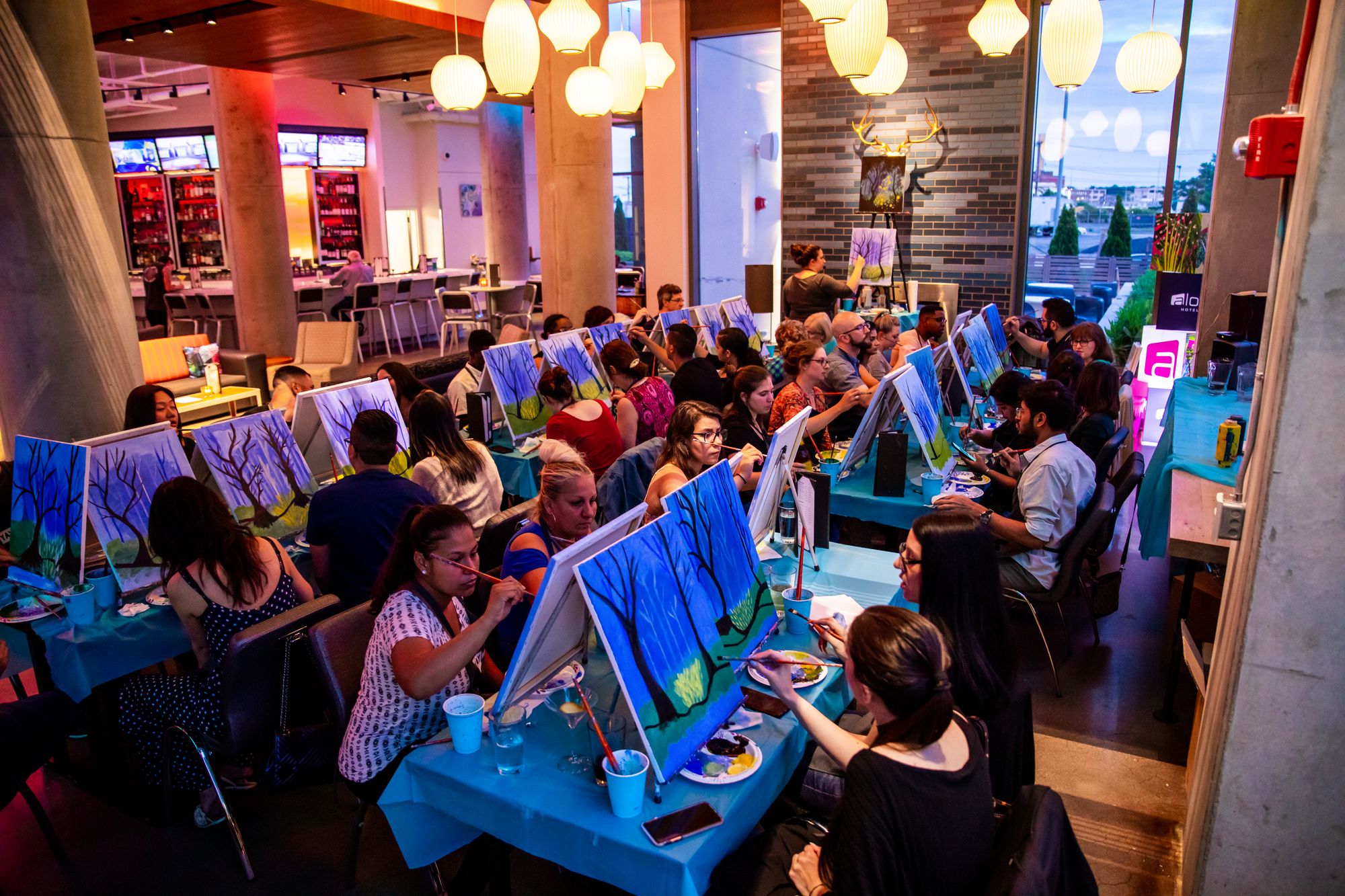 Paint Nite guests painting blue woodland scenes at tables inside a bar