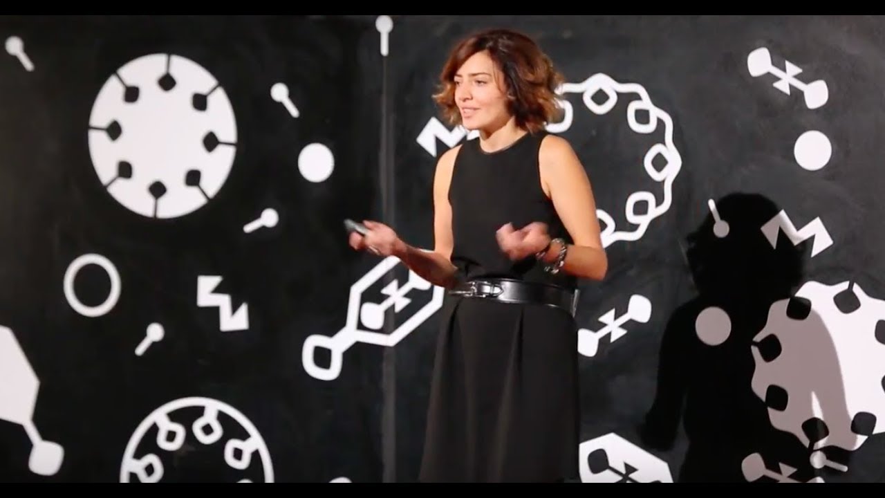 Yeva Hyusyan speaking on the TEDxYerevan stage, with white geometric designs behind her.