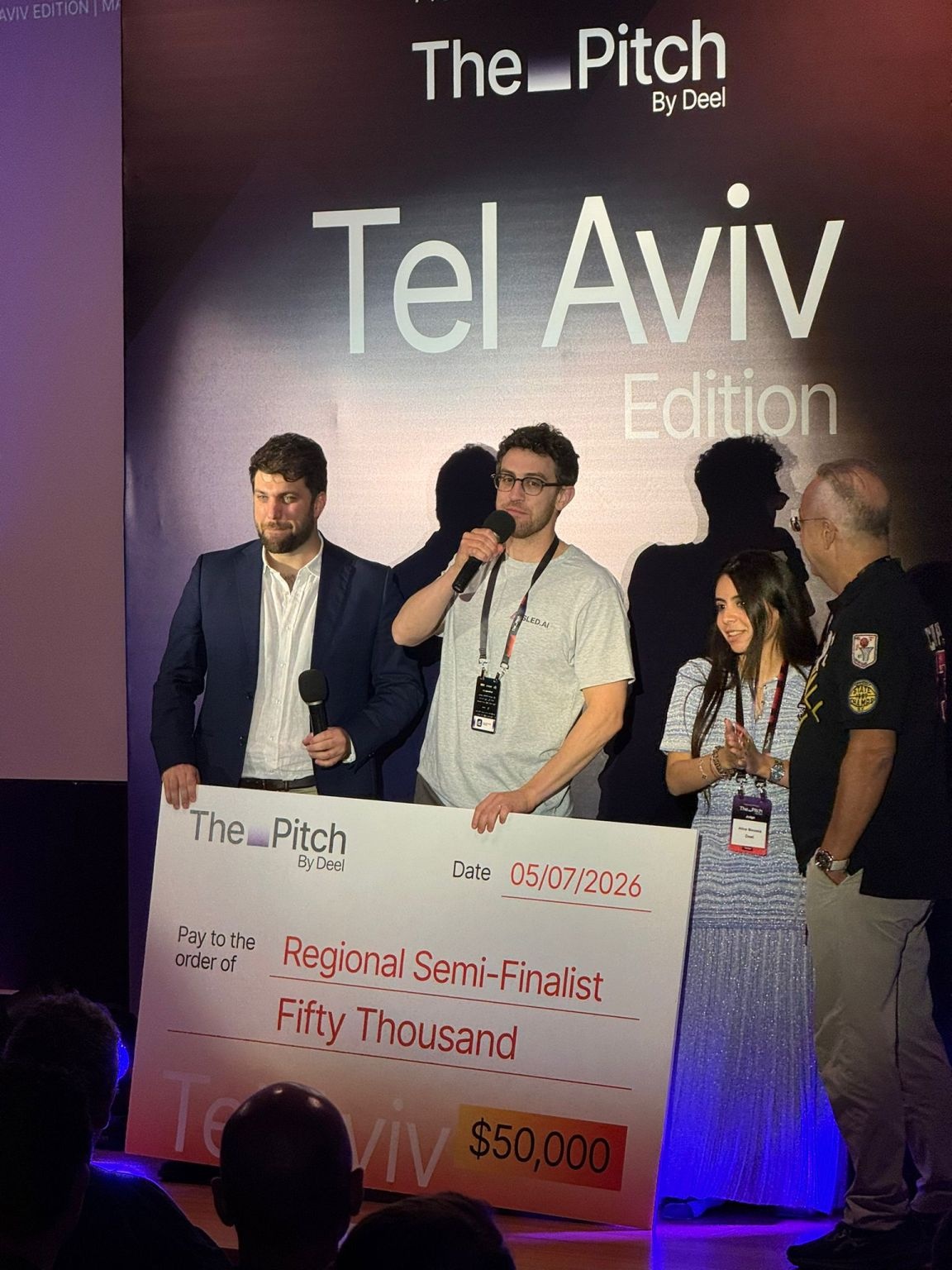 Yonatan Pistiner speaking into a microphone at The Pitch by Deel in Tel Aviv, beside a fifty-thousand-dollar competition check