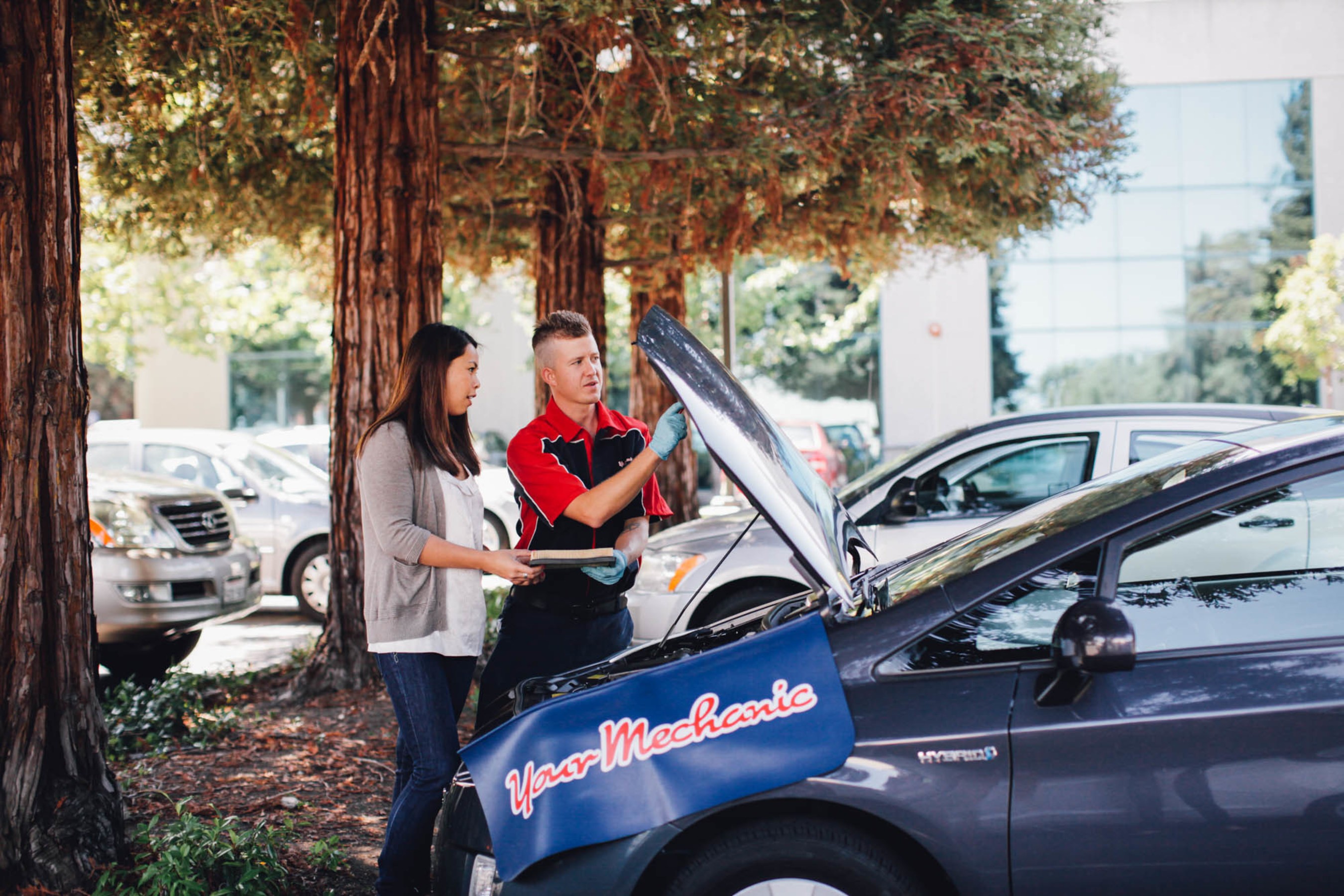 A YourMechanic technician discusses an open engine bay with a customer beside a parked car